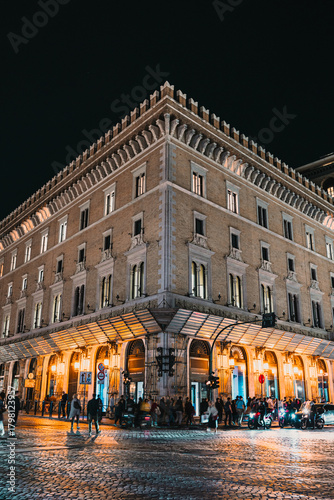 Exterior corner view of a large, well-lit historic building on a cobblestone street at night with people gathered near the ground floor.