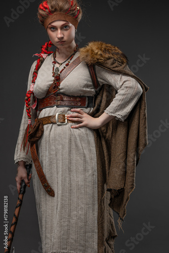 A young Viking woman, a combat heroine in a fantasy Nordic costume with a Scandinavian battle axe in her hands, a natural-colored dress and a cape with fur, photo on gray
