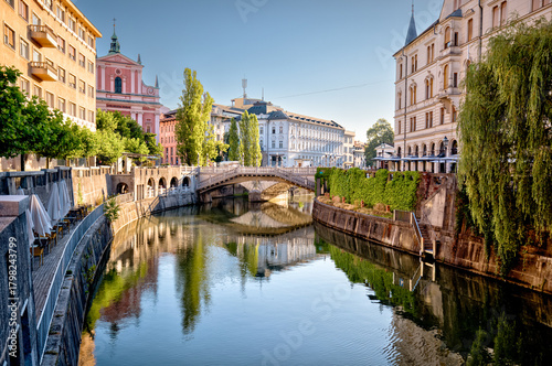 Fototapeta Naklejka Na Ścianę i Meble -  Ljubljana, Slovenia – July 14, 2025: A scenic view of the Ljubljanica River as it winds through the historic center of Ljubljana. Lined with trees, cafés, and charming riverside architecture.
