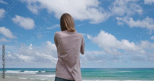 Woman stands on the beach, capturing the view of the ocean with their smartphone. The sun shines brightly as waves crash gently.