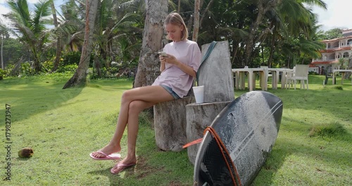 Woman sitting on a log by the beach while using her phone near a surfboard and palm trees. 