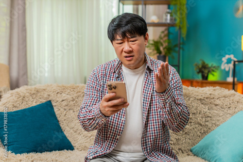 Фототапет Asian man sitting on home sofa reads bad fail news on smartphone, frowning, showing disappointed frustrated mood