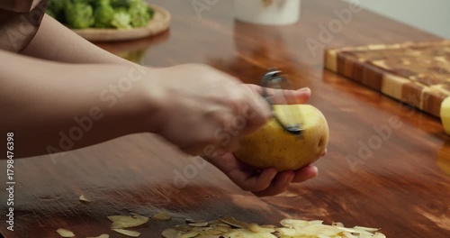 Close up of hands peeling fresh potato with peeler on wooden table, preparing ingredients for homemade vegan meal, cooking process, healthy food, rustic kitchen atmosphere, natural lifestyle