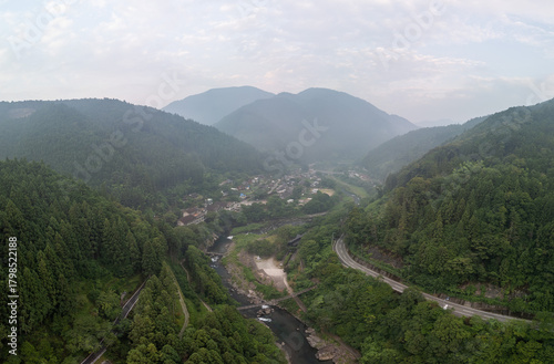 Aerial view of a lush valley with a winding river, surrounded by dense forests and mountains. A road snakes through the landscape, leading to a small town nestled among the trees. 