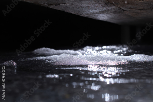 Water pooled beneath old crawlspace piping glistening surface with foam bubbles reflecting light in dark space