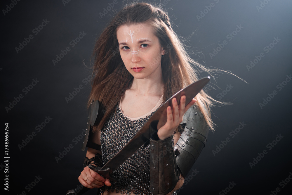 Naklejka premium Portrait of a young woman, fantasy warrior in armor with a sword in her hands isolated on a dark background