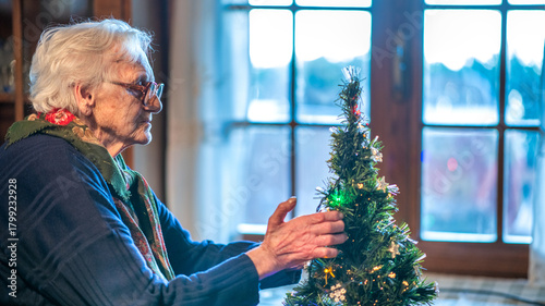 Elderly woman decorating a Christmas tree at home with colorful ornaments and warm lights