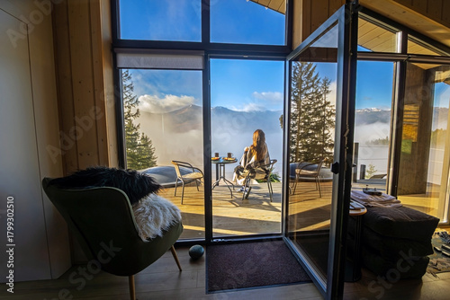 Woman enjoying mountain view from cozy cabin. A woman wrapped in a blanket sits on a terrace, drinking coffee and admiring a breathtaking Mountain View above the clouds.