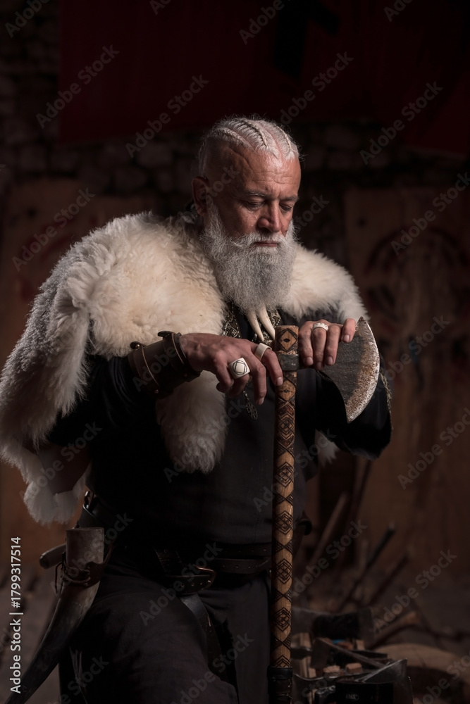 Fototapeta premium A rugged Viking man with a white beard, wearing traditional armor and a fur cloak, stands inside a stone hall, holding a sword with a contemplative expression, evoking strength and heritage.