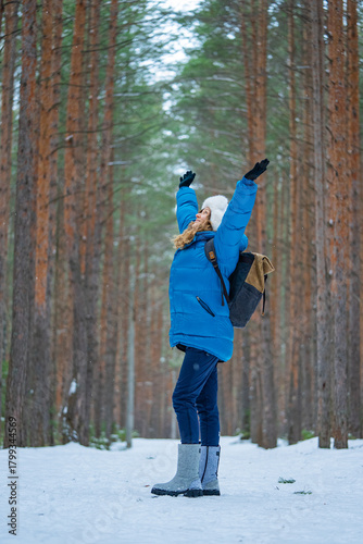 Woman in a blue jacket, white hat, and backpack standing in a snowy pine forest with arms raised, expressing excitement and freedom during a winter outdoor activity