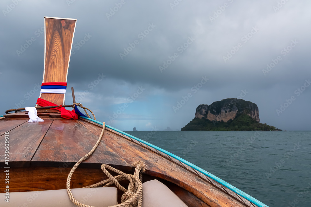 Obraz na plátně The wooden prow of a traditional Thai long-tail boat, decorated with a flag, sails through choppy sea towards distant island under an ominous, dark grey sky, right before a tropical storm in Krabi