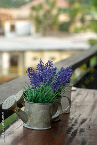 A rustic metal watering can pot filled with purple artificial flowers sitting on a wooden railing. A charming decorative touch adding a vintage feel to a balcony or cafe setting.