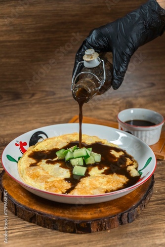 Slika na platnu Pempek with Vinegar Sauce: A close-up shot captures the delicious moment of a gloved hand pouring dark, rich vinegar sauce over a plate of pempek, garnished with fresh cucumber slices
