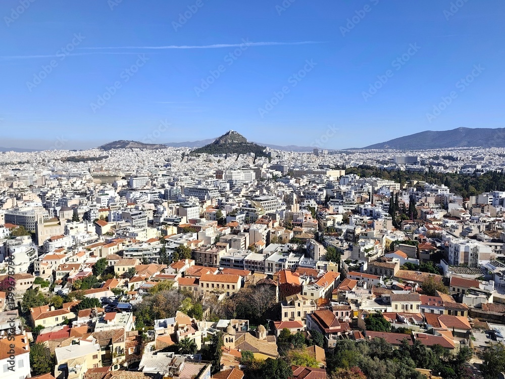 Fototapeta premium Panoramic view of Athens, Greece, under a vivid blue sky, seen from the Acropolis Hill summit, with the charming Anafiotika neighborhood below and Lycabettus Hill rising in the distance.