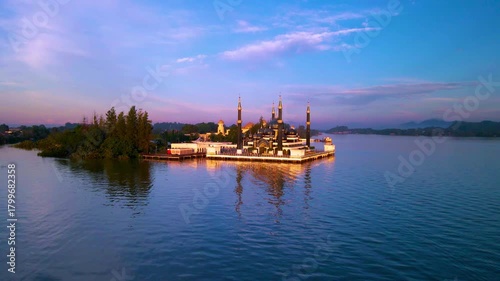 Aerial view of the Crystal Mosque at sunrise, with golden light reflecting on calm waters and clear blue skies around the iconic Islamic landmark.