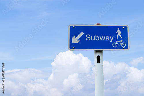 Signs dark blue rectangle aluminum with white letters go to subway, arrow and pedestrian icon with blue sky cloud white is background. Provides directions to underground metro station sign.