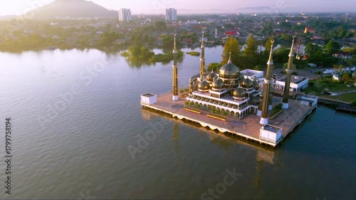 Aerial view of the Crystal Mosque at sunrise, with golden light reflecting on calm waters and clear blue skies around the iconic Islamic landmark.