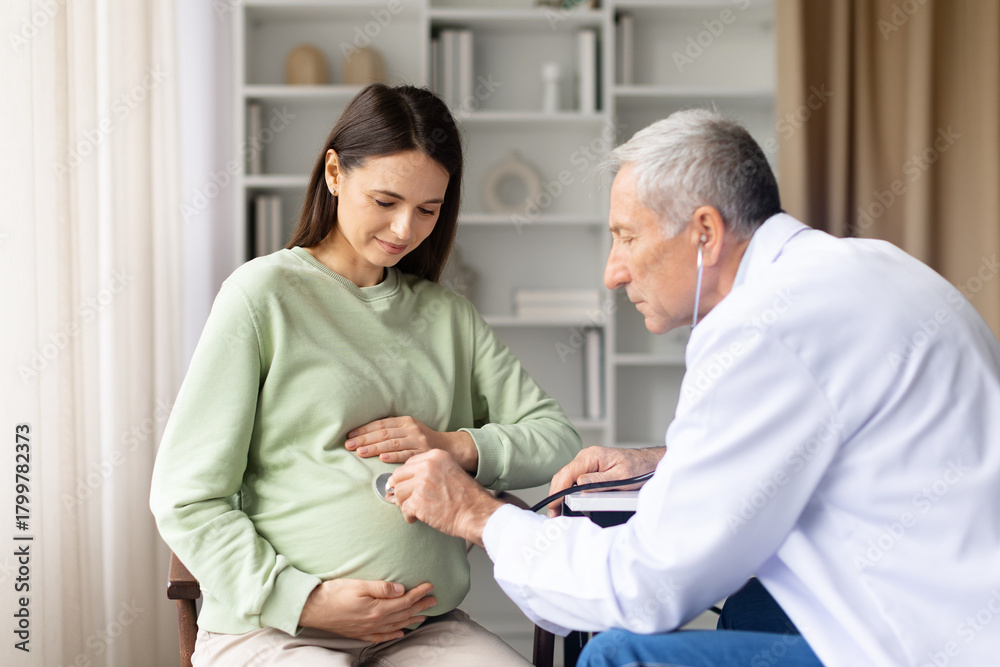Fototapeta premium Senior male doctor using stethoscope to examine pregnant womans belly during prenatal checkup in clinic, maternity healthcare and pregnancy care concept