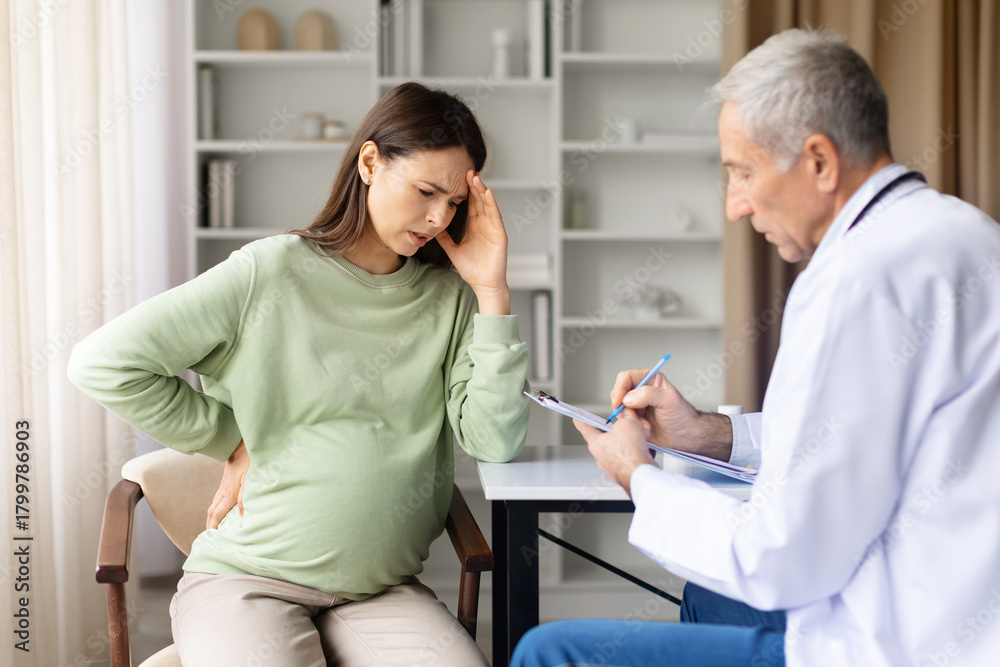 Fototapeta premium Pregnant woman suffering headache and back pain consulting senior male doctor in clinic office, pregnancy discomfort and prenatal healthcare concept