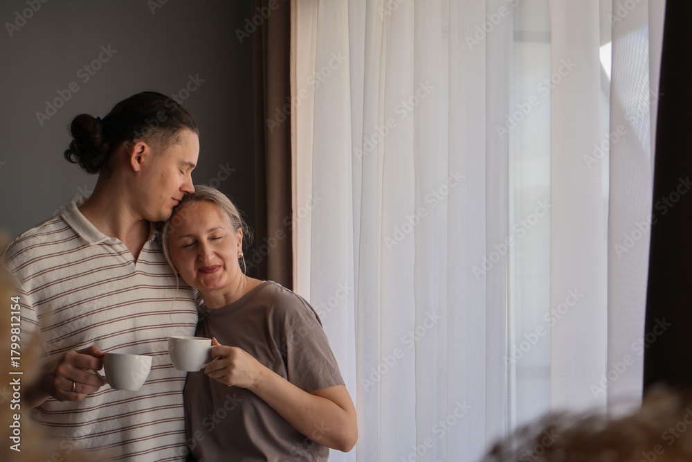 © Ina - Intimate morning moment: a loving mid-age couple enjoying coffee by the window, sharing a quiet connection and peaceful domestic routine.