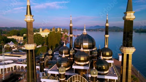 Aerial view of the Crystal Mosque at sunrise, with golden light reflecting on calm waters and clear blue skies around the iconic Islamic landmark.