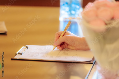 A close-up of a hand signing an official form on a clipboard using a gold pen, with a bouquet of pink roses partially visible in the foreground. Warm lighting suggests a formal or ceremonial setting.