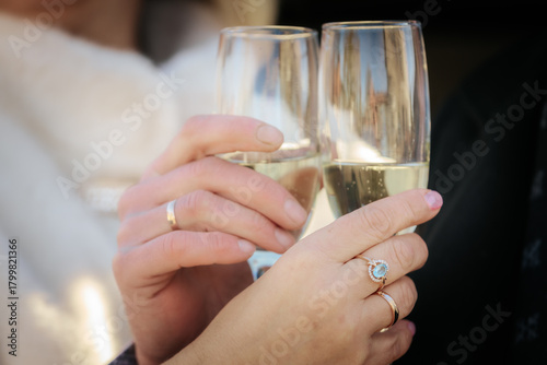 A romantic close-up of a couple clinking champagne glasses, both wearing wedding rings. The focus is on their hands and sparkling wine, suggesting celebration or engagement.
