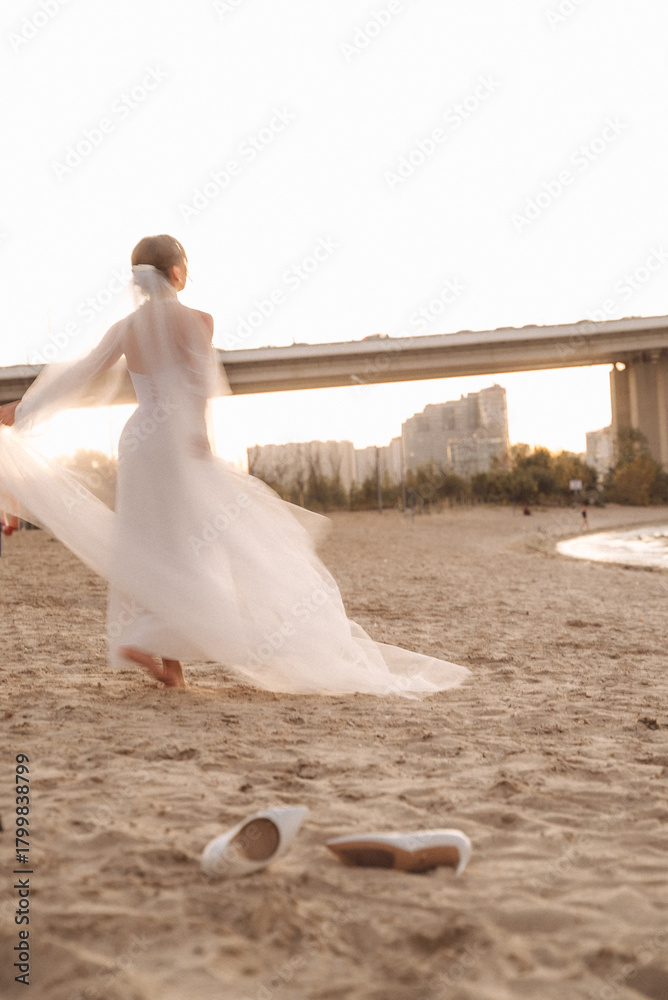 Naklejka premium Bride Dancing on a Sandy Beach at Sunset with Flowing Veil and Urban Bridge in the Background