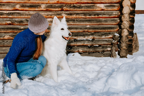 A woman in a knit hat and winter clothes sits beside a white dog in the snow, in front of a rustic wooden cabin. Peaceful outdoor winter moment with a loyal pet.