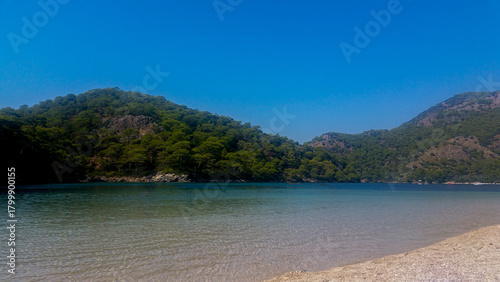 Fototapeta Naklejka Na Ścianę i Meble -  Sandy Beach and Calm Waters — Green Hills, Blue Sky, and Coastal Stillness in Oldöniz, Turkey