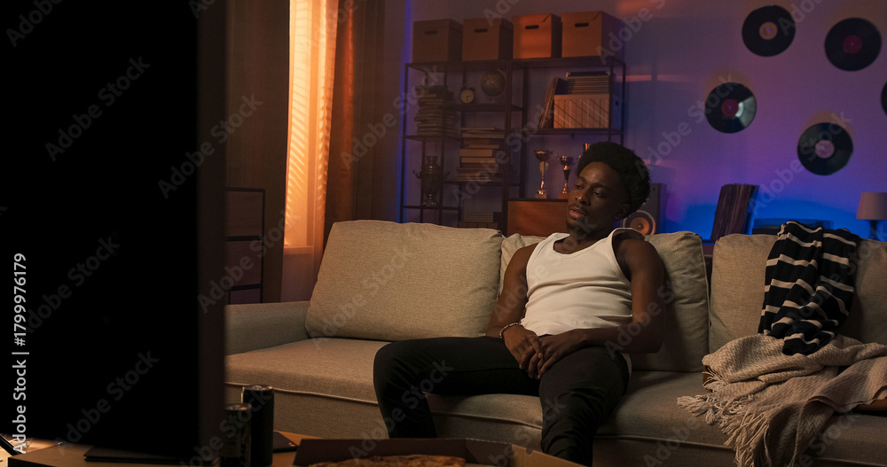 Obraz premium A pensive man of different nationalities dressed in a white T-shirt with straps sits on a sofa, quietly staring into the distance, showing signs of sadness and personal reflection.