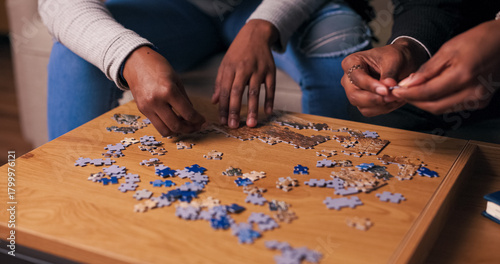 A close-up of the hands of two young people of multi-racial background working on a puzzle. They search for the right pieces and assemble the picture together on a wooden table.