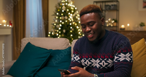 A young cheerful guy sits on the couch and talks on the phone. He communicates with girlfriend and smiles. The guy invites on date. A Christmas tree flickers in the room.