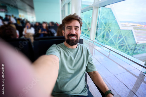 Smiling caucasian millennial man taking a selfie while waiting at an airport gate, seated by large glass windows. Bright terminal architecture conveys travel mood and anticipation.