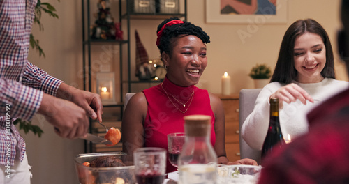 Sitting at the table and smiling during a friend's birthday celebration, are a girl with dark hair in a white sweater and a multi-ethnic girl in red clothes. A guy in a shirt cuts meat and puts it on