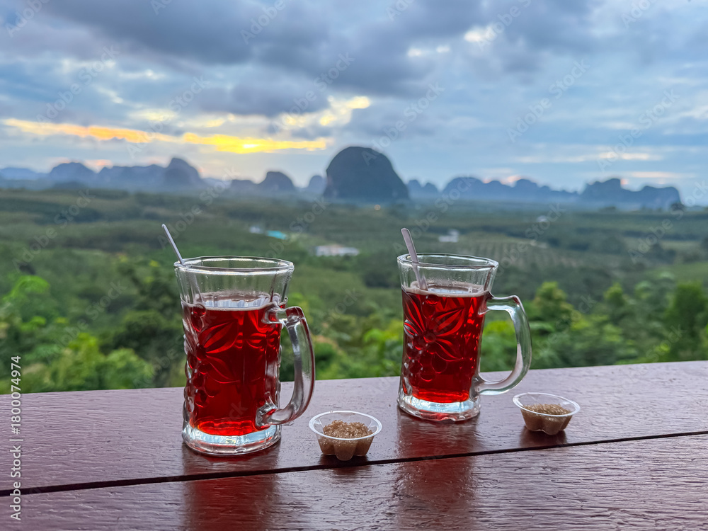 Naklejka premium Two glasses of red tea rest on a balcony railing, offering a romantic start to the day overlooking the stunning mountain landscape of Krabi, Thailand, during a beautiful sunrise.