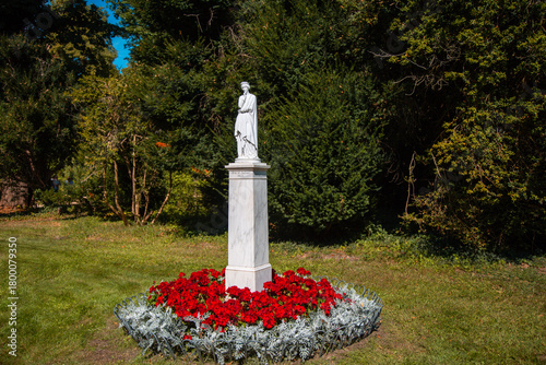 Eine weiße Statue auf einem Sockel umgeben von roten Blumen ind silbernen Laub