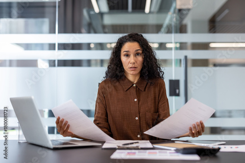 Stressed businesswoman sitting at a modern office desk, looking directly at the viewer with a concerned expression while holding papers, signifying workload, financial problems, and confusion
