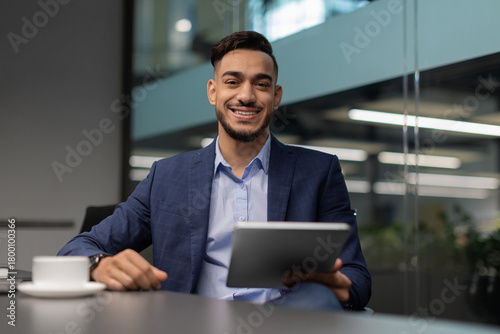 A cheerful middle eastern businessman in a neat suit sits at a table in a bright office. He is using a digital tablet to explore the latest business applications while smiling happily.