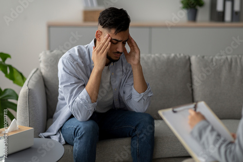 Φωτογραφία A stressed man sits on a couch, touching his temples in frustration