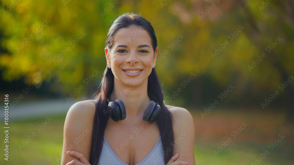 Obraz premium Happy young woman wearing headphones around her neck and sportswear, looking at the camera and smiling, feeling positive and active in a green park environment