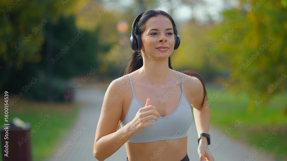 Fototapeta premium Woman jogging on a path in a park, wearing a sports bra and wireless headphones, with a smartwatch on her wrist while engaging in outdoor exercise and a healthy lifestyle