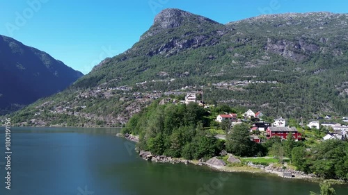 Drone flying over Norwegian fjords, showing city houses by the water, calm blue fjord surface, steep surrounding mountains, and cars moving along coastal roads.