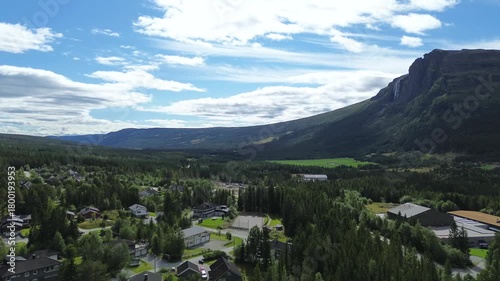 Drone flying over city houses in Norway, revealing a mountain ahead with a waterfall flowing down its side in a beautiful scenic landscape.