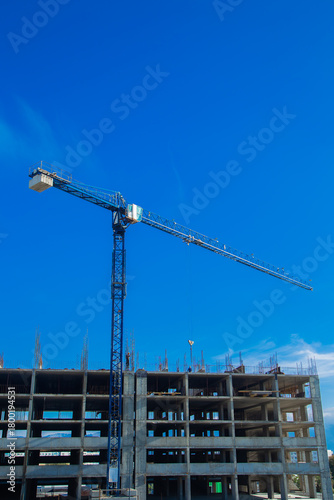 A tall construction crane on a construction site with a clear blue sky and a concrete structure illustrating the dynamic nature of urban development and construction work. Concept of construction