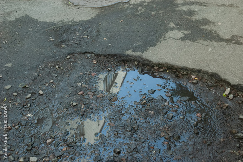 Broken asphalt with a pothole filled with gravel and a puddle reflecting a building.