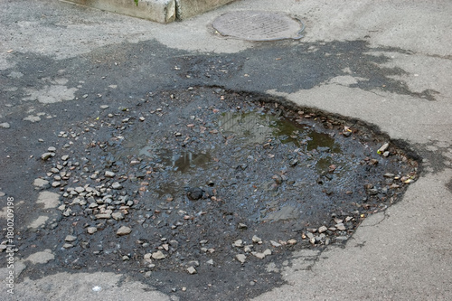 Broken asphalt with a pothole filled with gravel and a puddle reflecting a building.