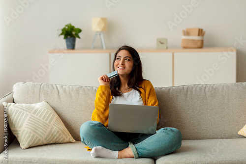 A pensive Arab lady sits comfortably on her sofa, holding a credit card and a laptop. She gazes thoughtfully into the distance as she considers her online purchases in a cozy living room setting.