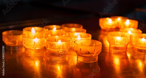 Group of amber-colored church candles burning on a reflective metal surface, creating a warm, peaceful glow in a dark indoor setting