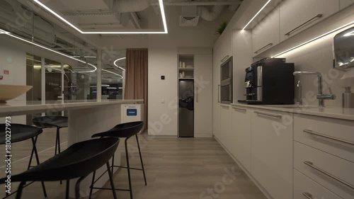 Modern office kitchen with sleek white cabinets, integrated appliances, bar stools, and LED ceiling lights, captured in a clean contemporary workspace environment during evening hours